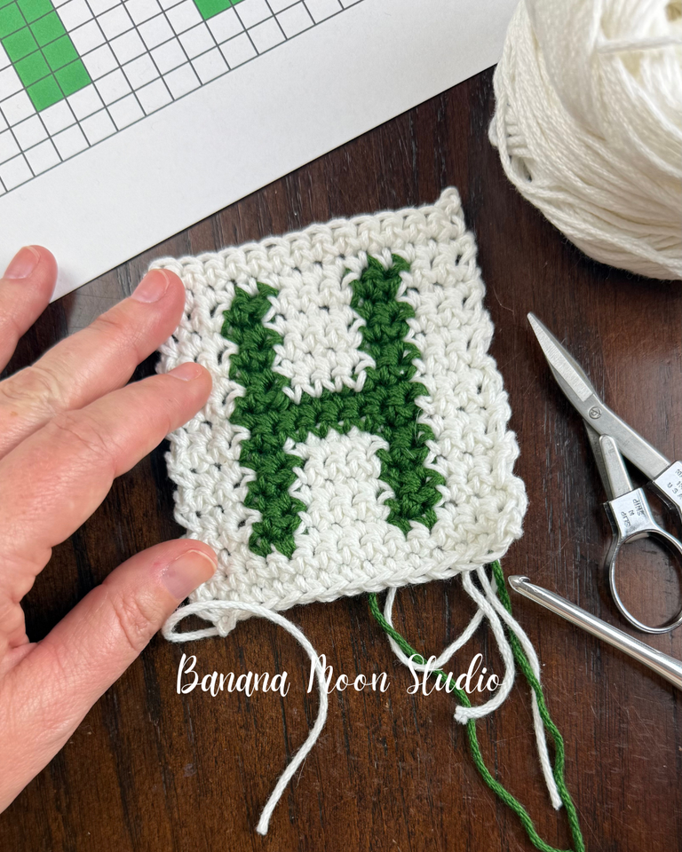 Small crochet piece in white with a green capital H stitched into it using crochet intarsia technique. Text on the image reads: banana moon studio.