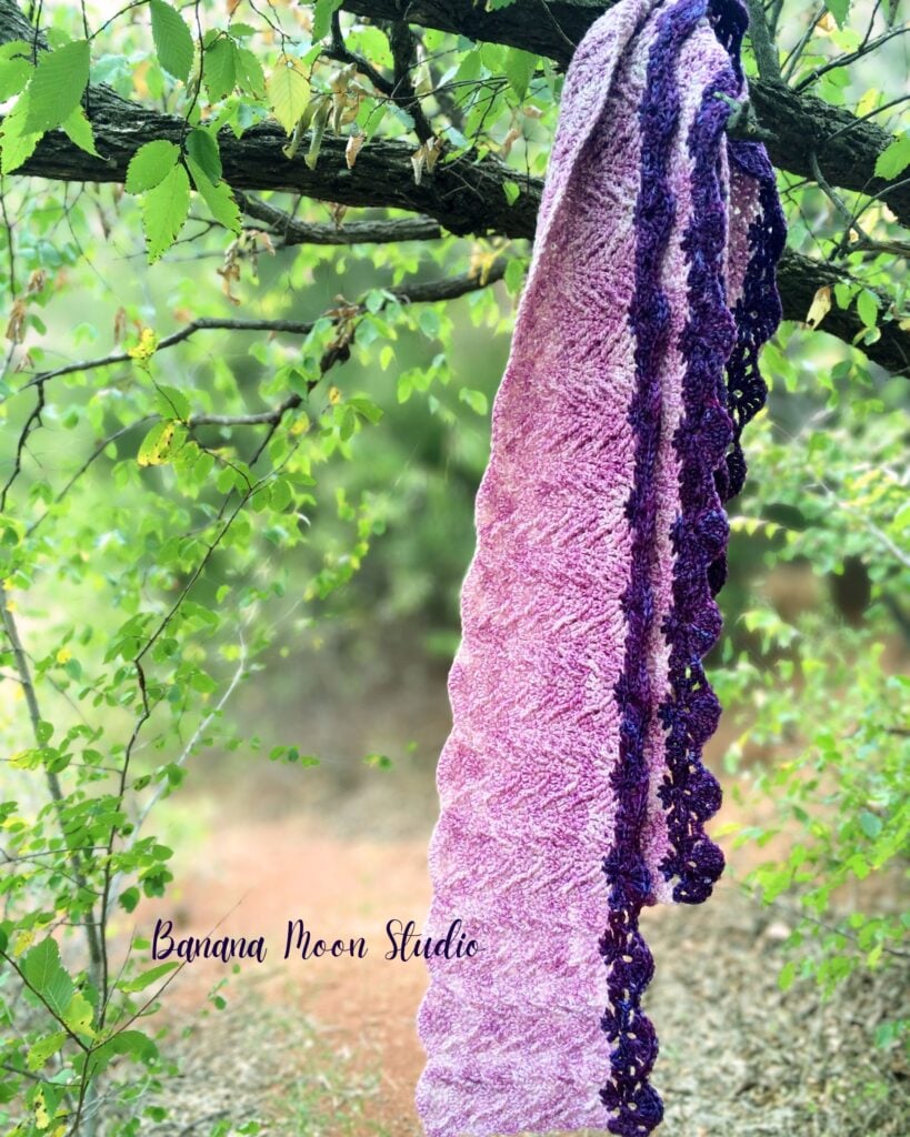 Purple women's crochet scarf pattern folded and hanging from a tree branch. Text on the image reads: Banana Moon Studio.