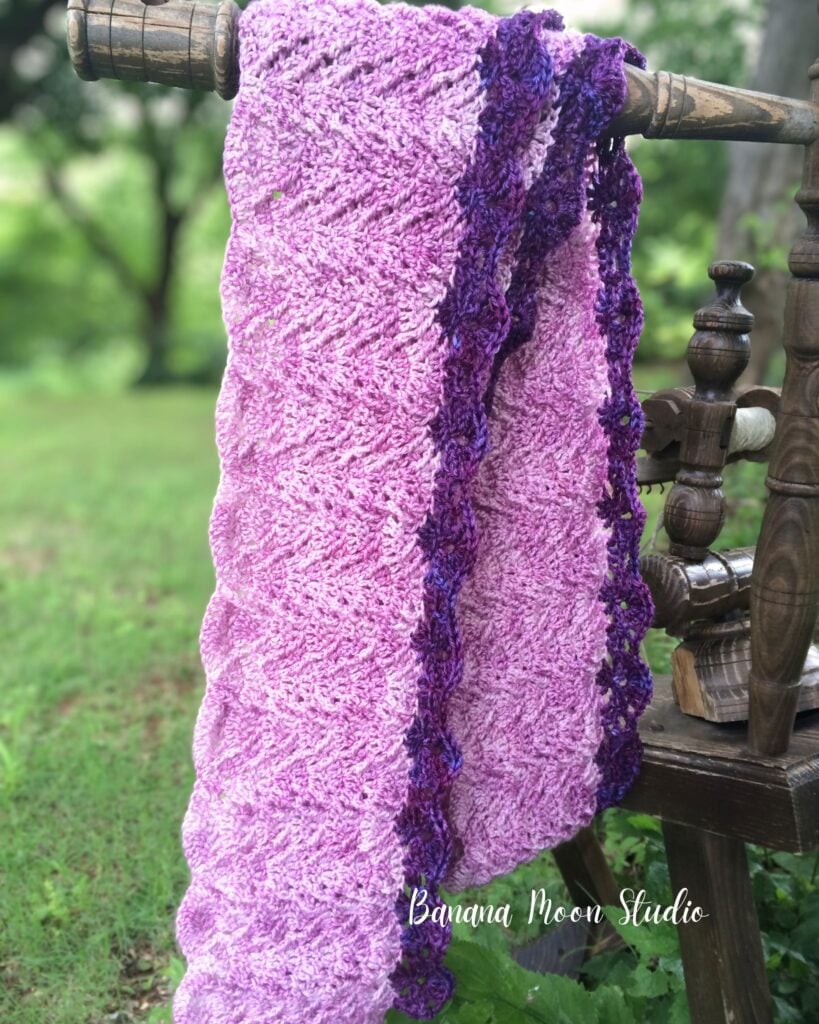 A purple textured crochet scarf folded over a wooden rod with woods in the background. Text on the image reads: Banana Moon Studio.