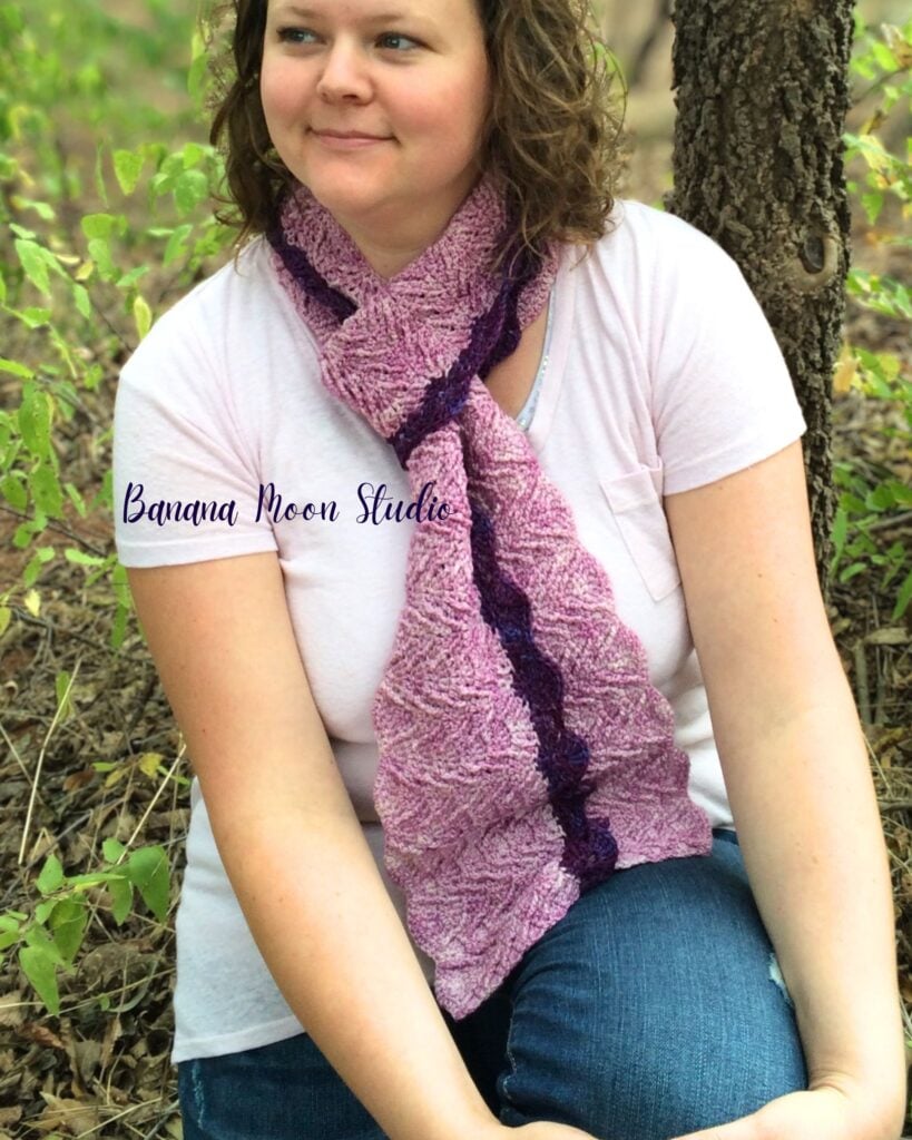 A woman kneeling in the woods wearing a textured crochet scarf. Text on the image reads: Banana Moon Studio.