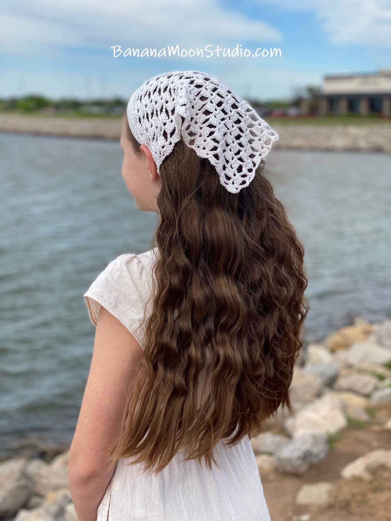 Girl standing near the water wearing a white lace hair kerchief and white jumpsuit. Marietta Kerchief free lacy crochet kerchief pattern from Banana Moon Studio.