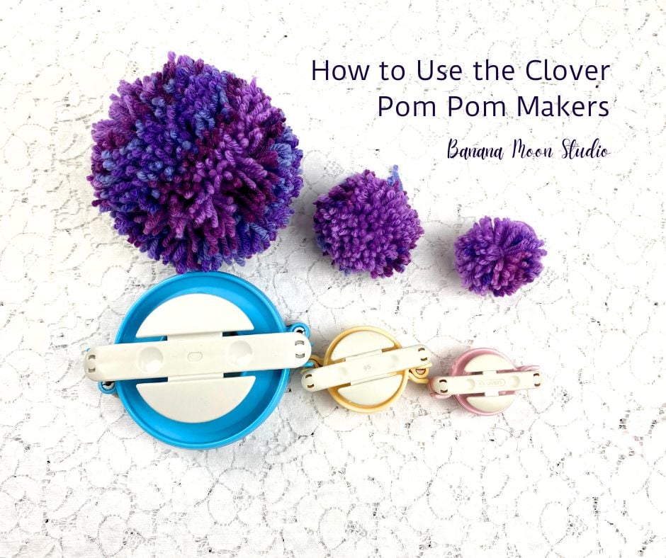 Three purple pom poms made of yarn lay on a white lace surface along with three pom pom makers. Text on the image reads: How to use the clover pom pom makers. Banana Moon studio.