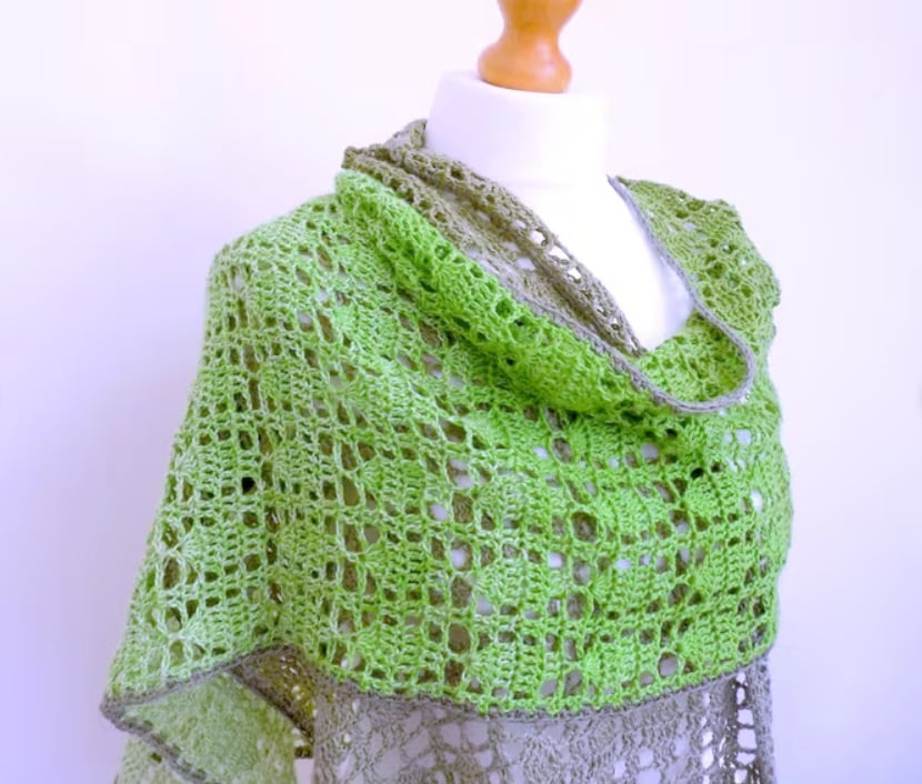 A crochet lace shawl with leaf shaped stitched throughout in a grid is wrapped around the shoulders of a white mannequin. A light purple wall is in the background.