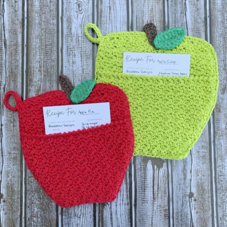 Red and green apple-shaped crochet hot pads each with a pocket. A recipe card is in each pocket. The hot pads sit on a wooden surface. 