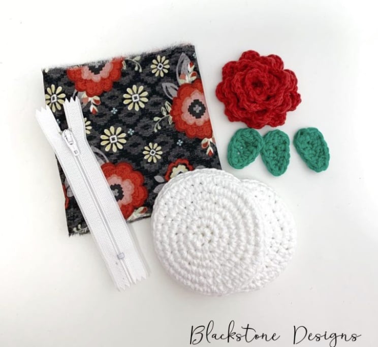 Printed material, a white zipper, two crochet circles, a red crochet rose, and three green crochet leaves all sitting together on a white surface. Text on the image reads: Blackstone Designs.