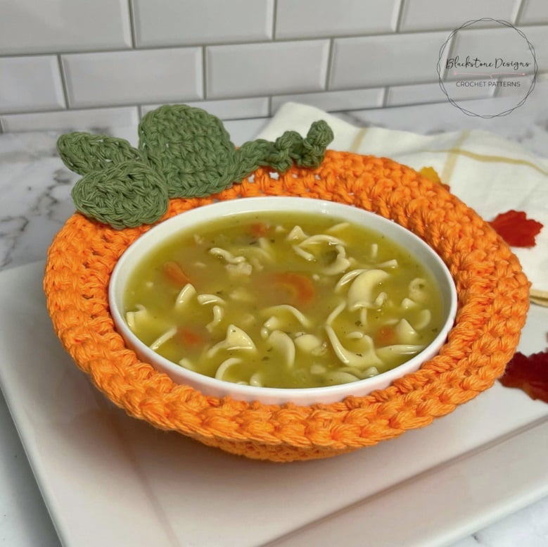 A white bowl of noodle and vegetable soup sits in an orange crochet bowl cozy made to look like a pumpkin with leaves, stem, and a curly vine at the top. It sits on top of a book on a marble countertop with a white tile backsplash. Text and logo on the image reads: Blackstone Designs Crochet patterns.