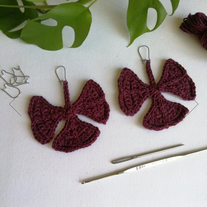 Small crochet oxalis leaves attached to earring hooks laying on a white surface with a crochet hook, tapestry needle, additional earring hooks, and green leaves. 
