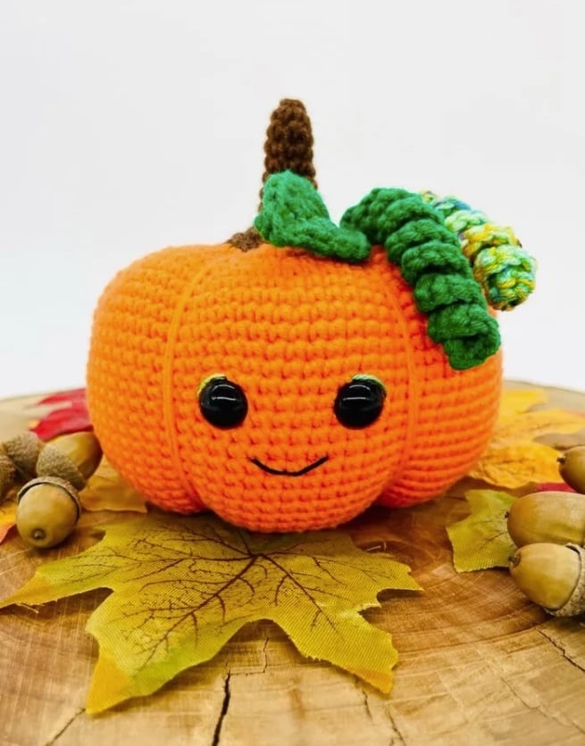 Bright orange crochet pumpkin with crochet stem, leaf, and curling vines. The pumpkin has plastic eyes and an embroidered mouth. It sits on a round wooden piece with leaves and acorns.