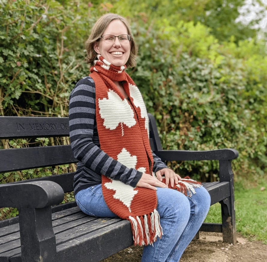 A woman its on a black park bench in front of a green hedge. She wears a wide crochet scarf in burnt orange with ecru leaves worked into it with intarsia.