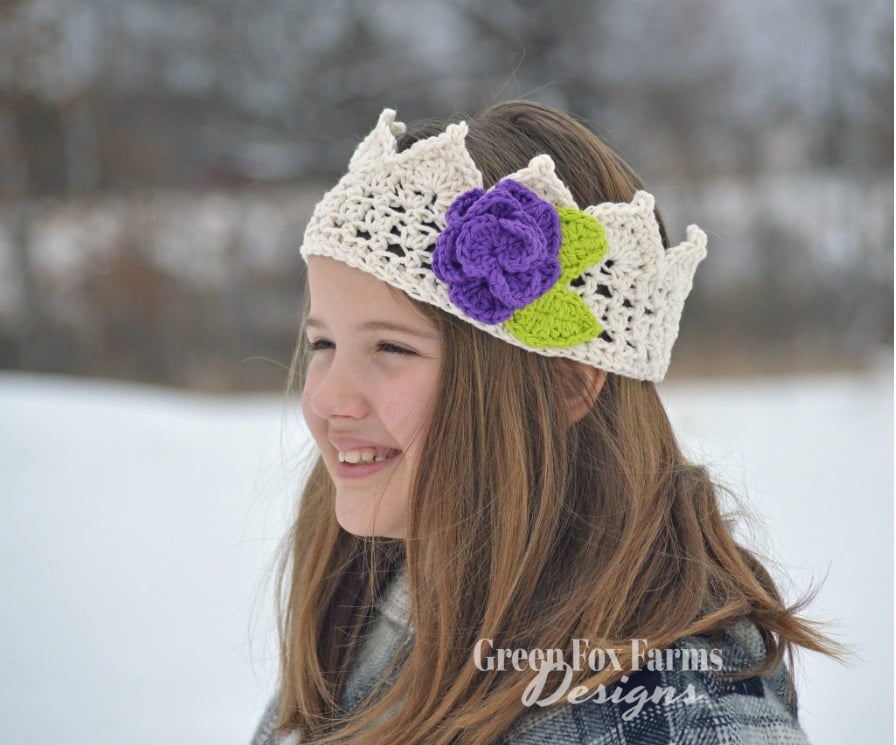 A child stands outside in the snow wearing a white crochet crown adorned with a purple crochet flower and two crochet leaves. Text on the image reads: Green Fox Farms Designs.