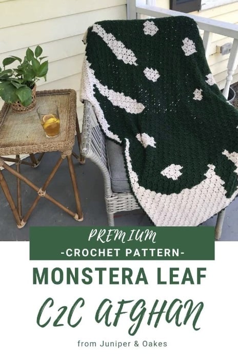 Crochet C2C blanket with a large, dark green monstera leaf stitched into a white background. The blanket lays draped over a whicker chair on a patio next to a table with a drink and potted plant on it. White and dark green panels with text read: Premium crochet pattern. Monstera leaf C2C afghan from Juniper & Oakes.