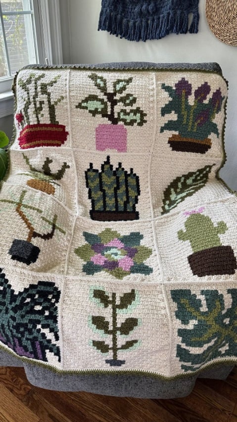 C2C crochet blanket with an ecru background. It is made of squares and each square has a plant or a leaf on it. The blanket is draped over an armchair with a window and white wall in the background.