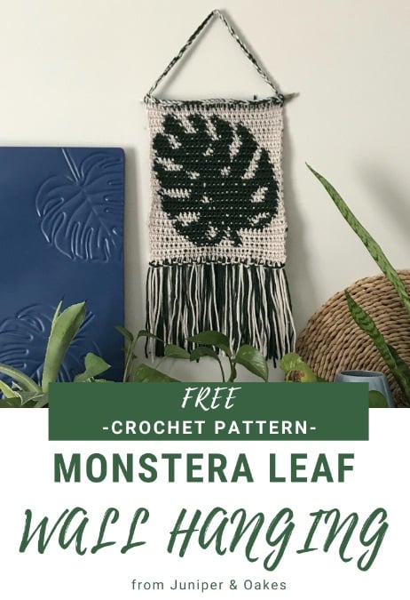 Crochet wall hanging with a monstera leaf crocheted in intarsia in an ecru background. The bottom edge is finished with fringe and the top is suspended from a stick and a string to hang it. White and green panels with text read: Free crochet pattern. Monstera leaf wall hanging from Juniper & Oakes.