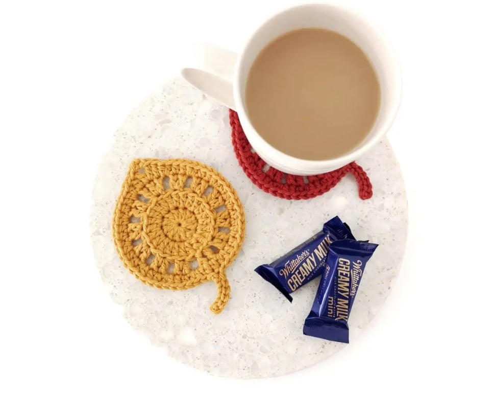 Yellow and red crochet leaves function as coasters and sit on an off white circle. A cup of coffee sits atop the red leaf coaster. Two small pieces of candy sit with them.
