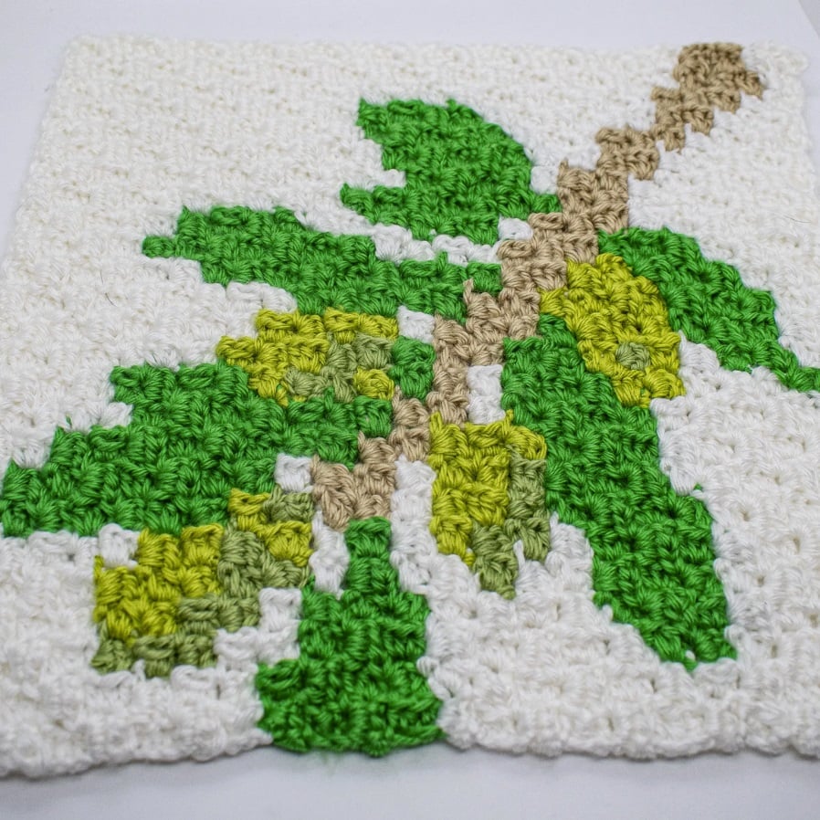 Close up photo of a C2C crochet square depicting the branch of an olive tree with leaves in two shades of green and a pale brown branch.