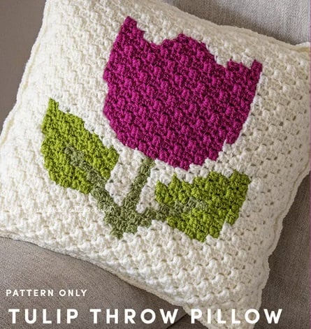 A C2C crochet pillow sits on a sofa. It has an ecru background with a fuchsia tulip and green stem and leaves on it. Text on the image reads: Pattern only. Tulip throw pillow.
