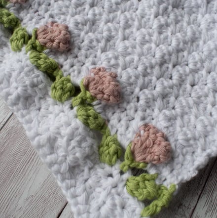 The edge of a crochet hand towel. The background of the towel is white with a border of dusty pink tulips with green leaves and stems.