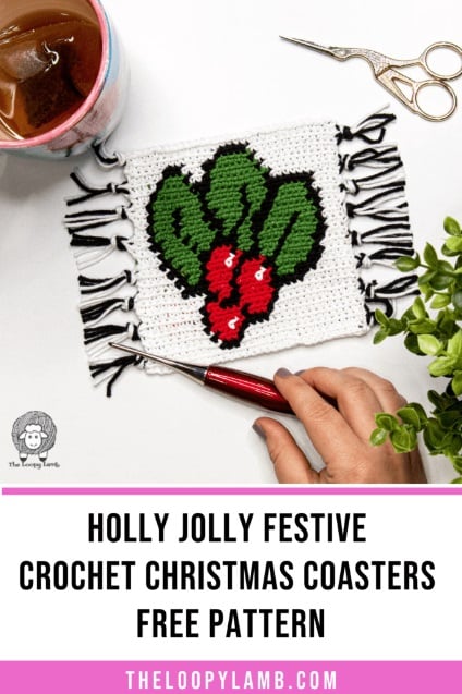 A crochet mug rug has a white background with holly leaves and berries on it outlined in black. The sides of the coaster are finished with white and black fringe. It sits on a white surface with a cup of tea, scissors, a plant, and a hand holding a crochet hook. Text and logo on the image reads: The Loopy Lamb. White and pink panels with text read: Holly jolly festive crochet Christmas coasters free pattern. TheLoopyLamb.com.
