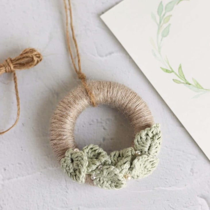 A small wreath wrapped in pale brown yarn with tiny pale green leaves and small pearl beads decorating the bottom of it. It has a length of twine at the top for a hanging loop and stis on a white surface with more twine and a piece of paper printed with a wreath.