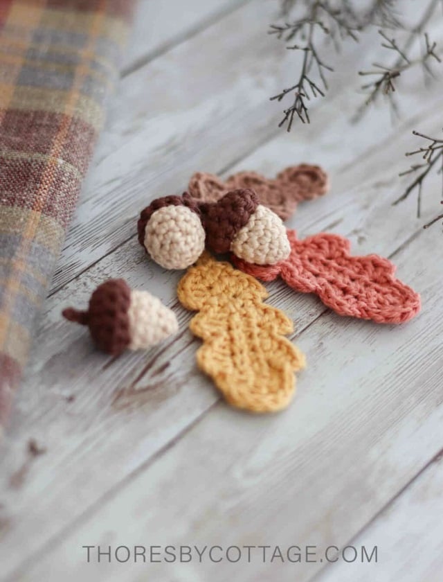 Crochet leaves and crochet acorns arranged in a small group on a wooden surface with plaid material and evergreen branches in the background. Text on the image reads: Thoresbycottage.com.