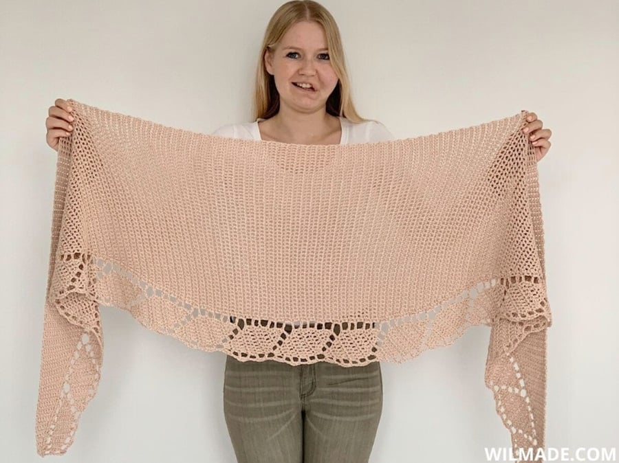 A woman stands against a white wall holding up a peach colored crochet shawl with a lace edging containing leaf shapes. Text on the image reads: Wilmade.com.