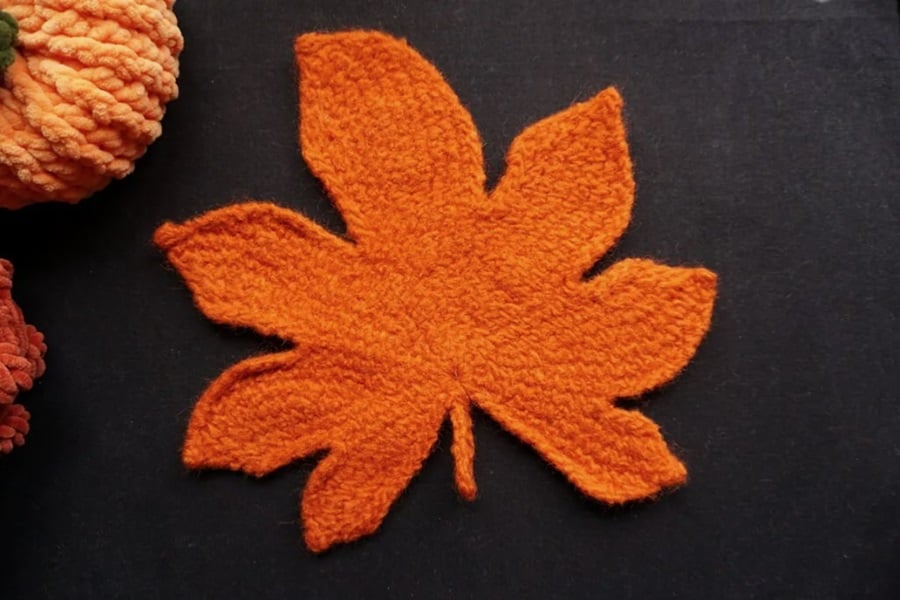 Pumpkin orange felted Tunisian crochet maple leaf on a black surface with additional crochet pieces in the background.