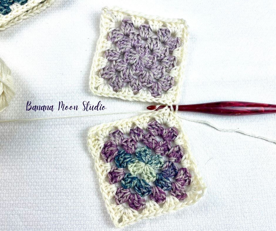 Two granny squares on a white surface with a red crochet hook and matching skein of yarn. The crochet hook is in the process of joining the two squares together. Text on the image reads: Banana Moon Studio.