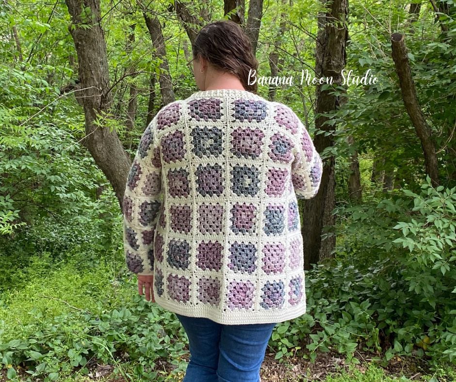 A woman standing in the woods wearing a granny square cardigan in cream and shades of purple and green over blue jeans. Her back is to the camera. Text on the image reads: Banana Moon Studio.