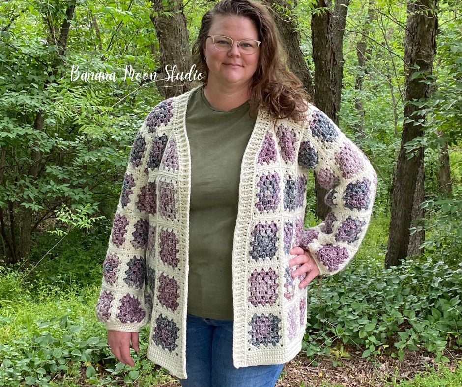 A woman standing in the woods wearing a granny square cardigan in cream and shades of purple and green over a green t-shirt and blue jeans. Text on the image reads: Banana Moon Studio.