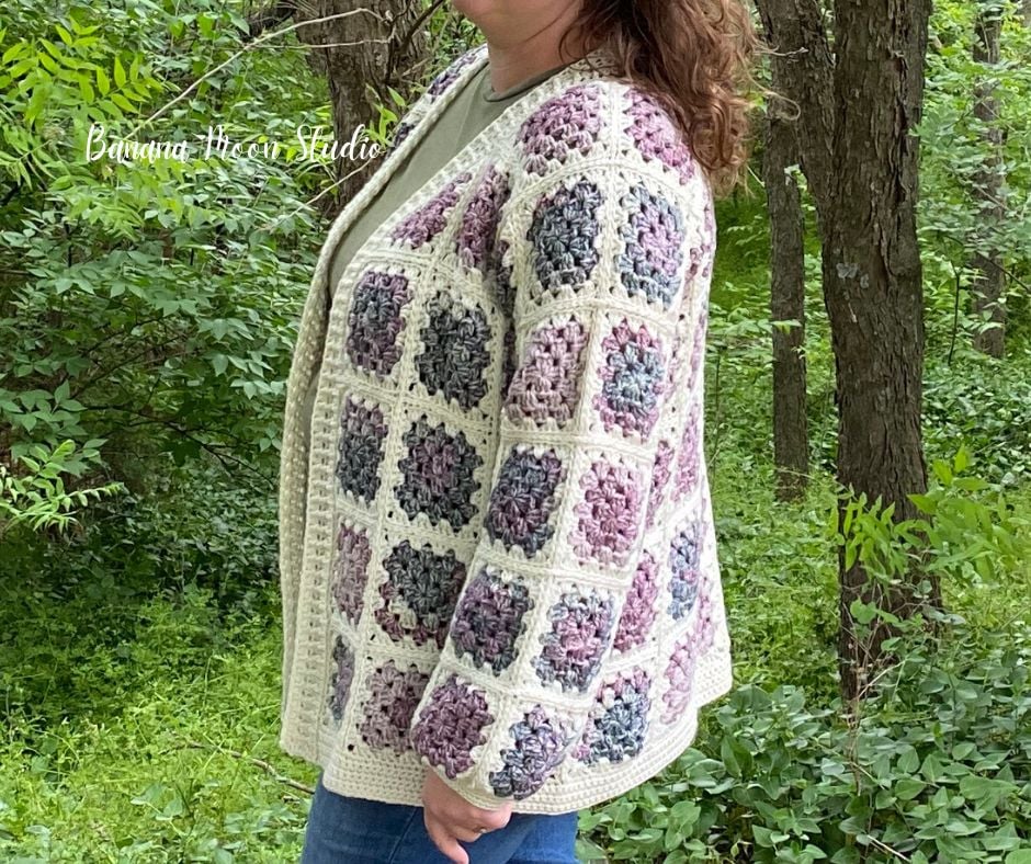 A woman standing in the woods wearing a granny square cardigan in cream and shades of purple and green over a green t-shirt and blue jeans. Text on the image reads: Banana Moon Studio.