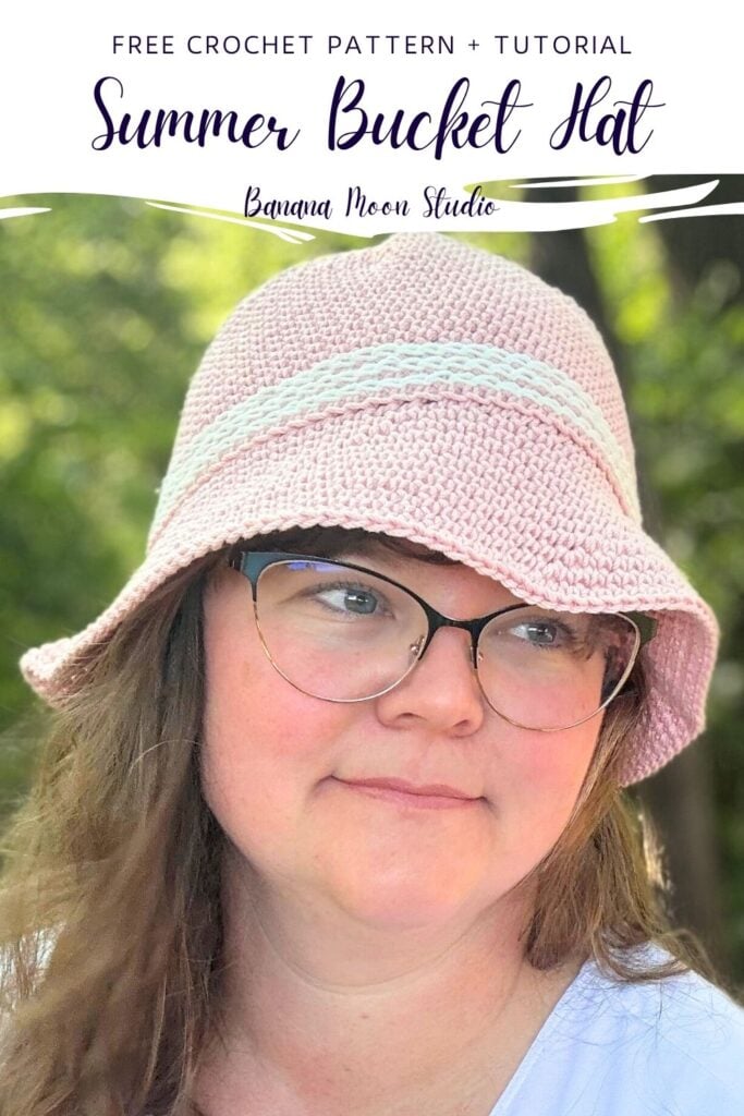 A woman in the woods wearing a pale pink crochet bucket hat with white trim. A white panel with text reads: Free crochet pattern + tutorial; summer bucket hat; banana moon studio.