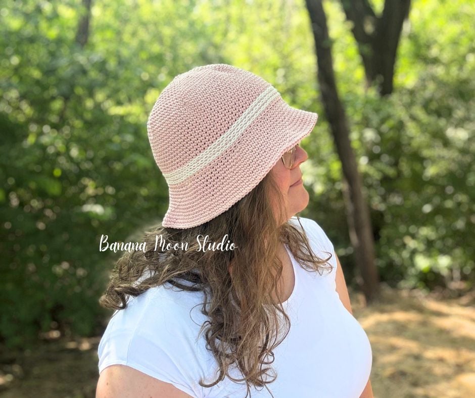 A woman in the woods, facing to the side, wearing a pale pink crochet bucket hat with shite trim. Text on the image reads: Banana Moon Studio.