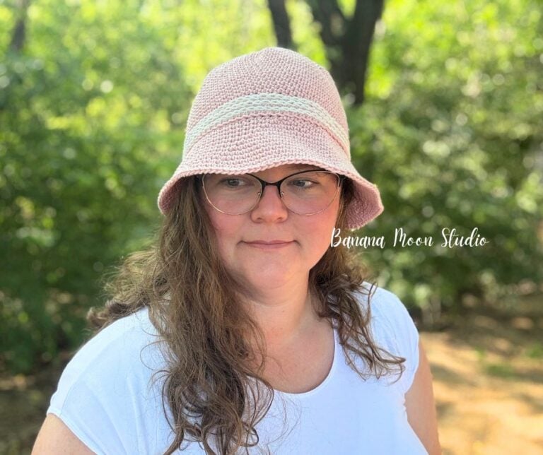 A woman in the woods wearing a pale pink crochet bucket hat with white trim. Text on the image reads: Banana Moon Studio.