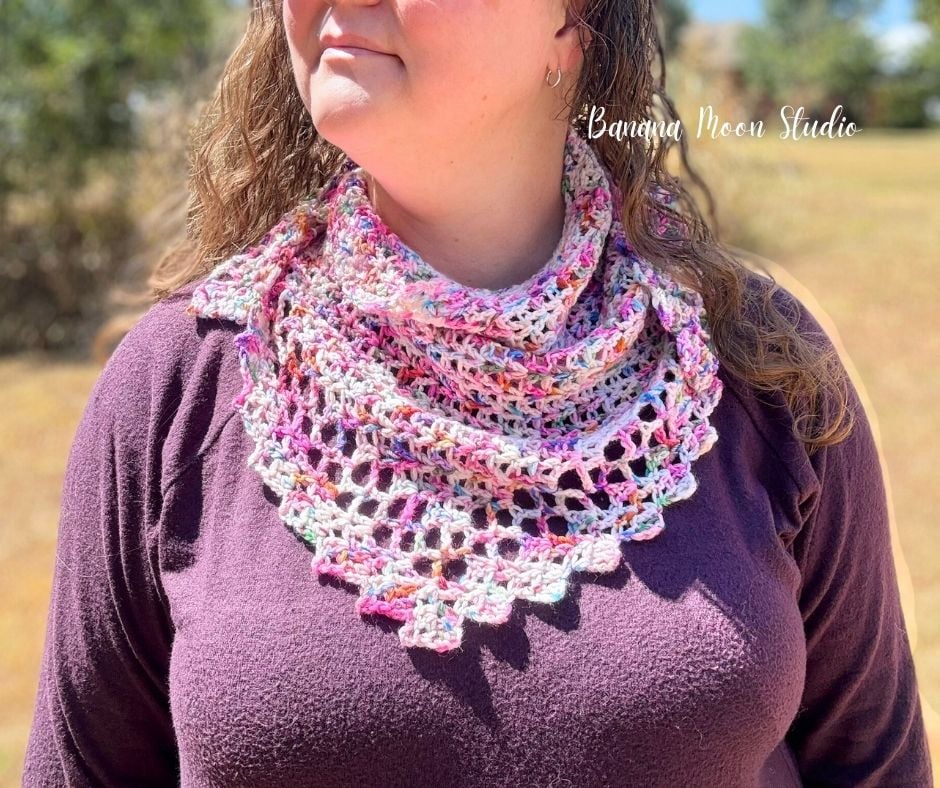 A woman stands outside wearing a crochet triangle scarf pattern with a lace edging over a sweater. Text on the image reads: Banana Moon Studio.
