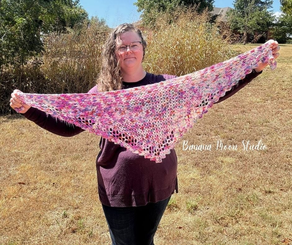 A woman stands outside holding a crochet triangle scarf out in front of her stretched out to show it. Text on the image reads: Banana Moon Studio.