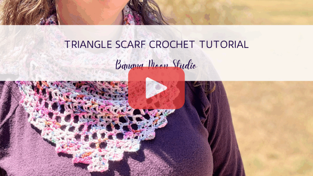 Close up photo of a woman wearing a triangular crochet scarf with a lace edging over a purple sweater. A banner with text reads: Triangle scarf crochet tutorial, banana moon studio. A red and white play button is centered over the image.