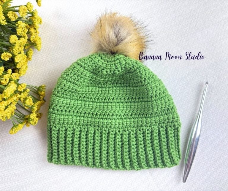 A crochet chemo cap pattern in lime green lying flat on a white surface with a crochet hook and yellow flowers. Text on the image reads: Banana Moon Studio.