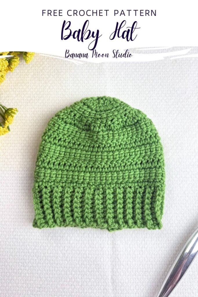 A lime green crochet baby hat lying flat on a white surface along with a silver crochet hook and yellow flowers. A panel with text reads: Free crochet pattern, baby hat, banana moon studio.