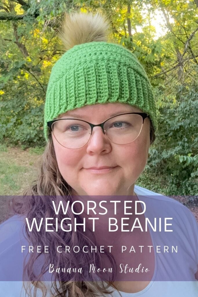 A woman standing outside wearing a lime green crochet beanie with a faux fur pom. A banner with text reads: Worsted weight beanie free crochet pattern, banana moon studio.