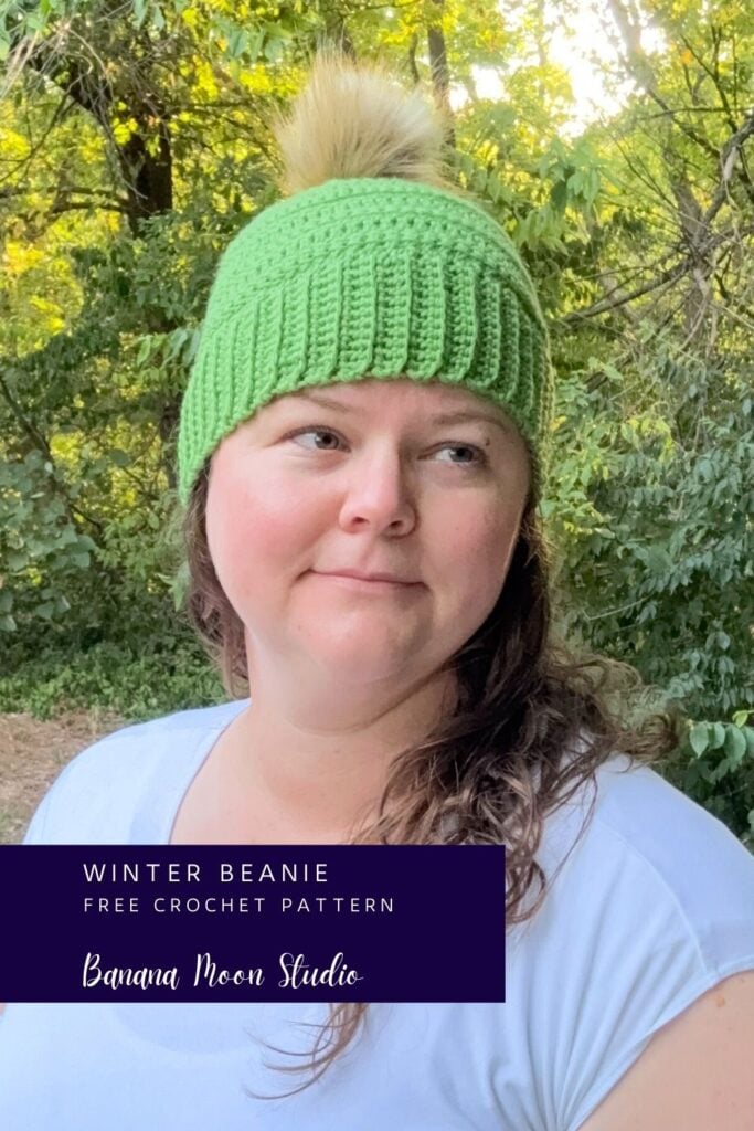 A woman standing outside wearing a lime green crochet beanie with a faux fur pom. A banner with text reads: winter beanie free crochet pattern, banana moon studio.