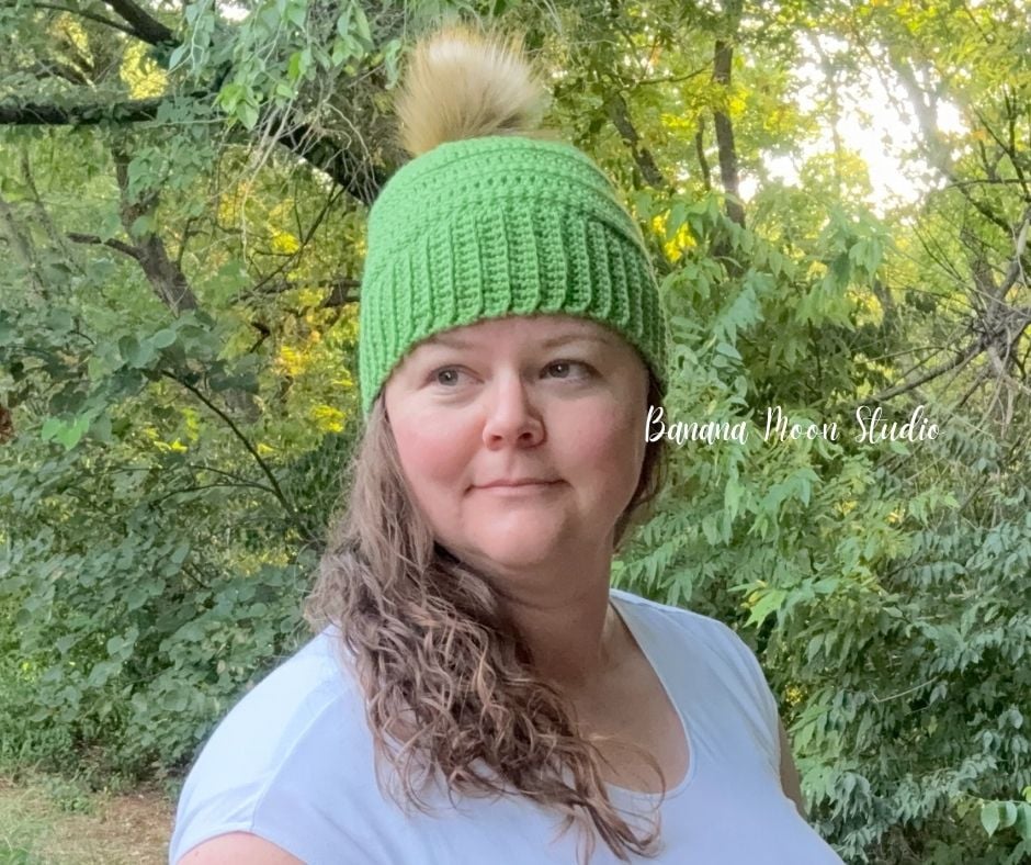 A woman in the woods wearing a lime green crochet beanie with a faux fur pom on top. Text on the image reads: Banana Moon Studio.