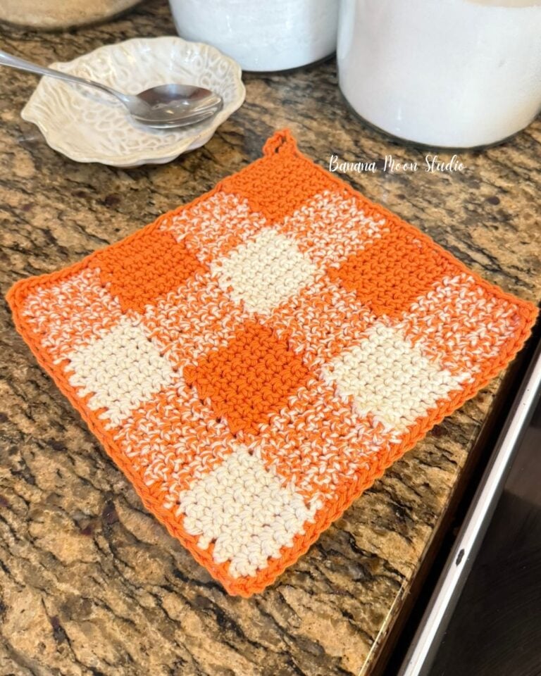 Orange and white crochet checkered pot holder laying on a granite countertop. Text on the image reads: Banana Moon Studio.