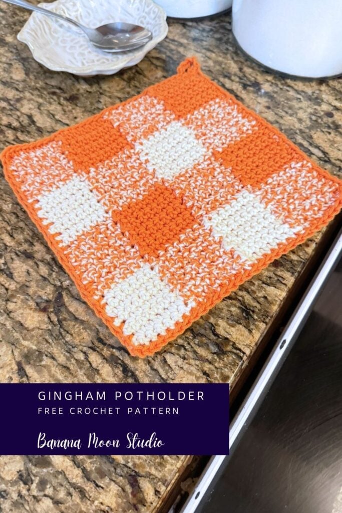 Orange and white gingham crochet pot holder lying flat on a granite countertop. A panel with text reads: gingham potholder free crochet pattern, banana moon studio.