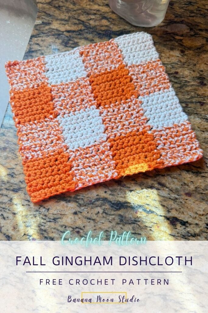Orange and white gingham crochet dishcloth lying flat on a granite countertop next to a sink. A panel with text reads: Fall gingham dishcloth free crochet pattern, banana moon studio.