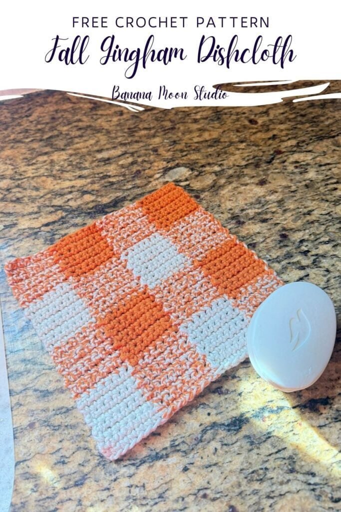 Orange and white gingham crochet dishcloth and a bar of soap sitting on a granite countertop. A panel with text reads: Free crochet pattern, fall gingham dishcloth, banana moon studio.