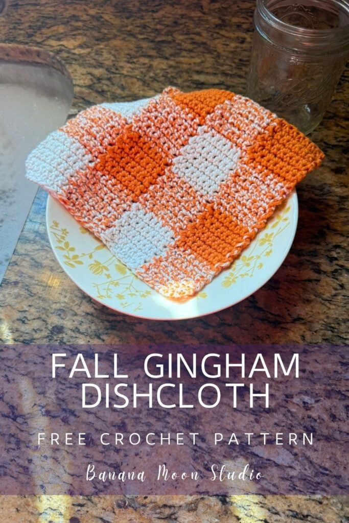 Orange and white gingham crochet dishcloth laying in a bowl on a granite countertop next to a sink. A panel with text reads: Fall gingham dishcloth free crochet pattern, banana moon studio.
