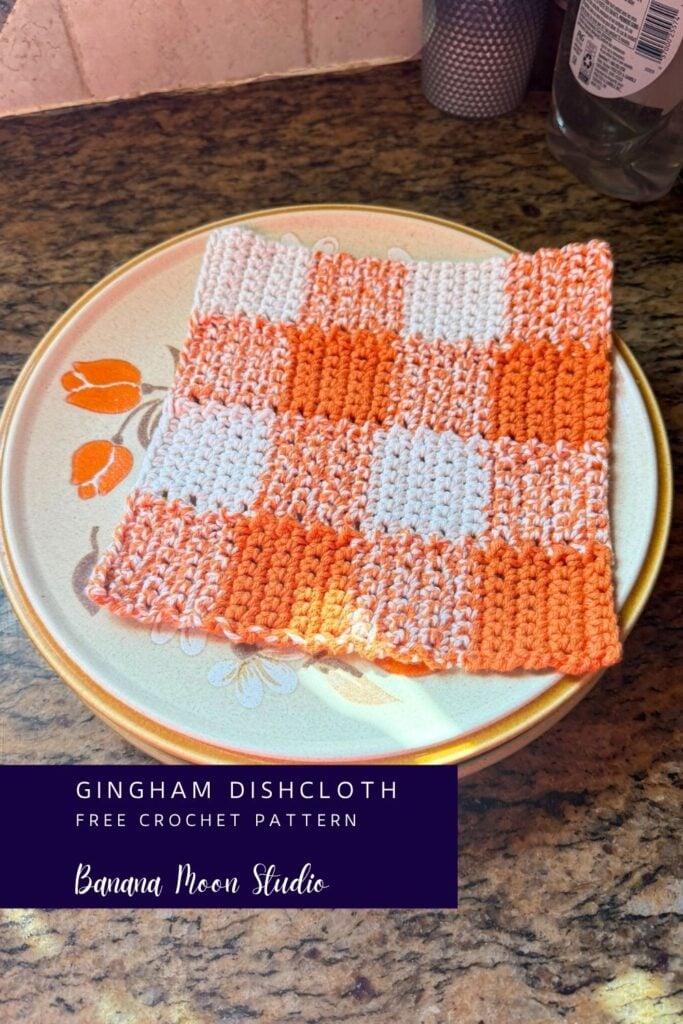 Orange and white crochet gingham dishcloth pattern laying flat on a ceramic dinner plate on a granite countertop. A panel with text reads: Gingham dishcloth free crochet pattern, banana moon studio.