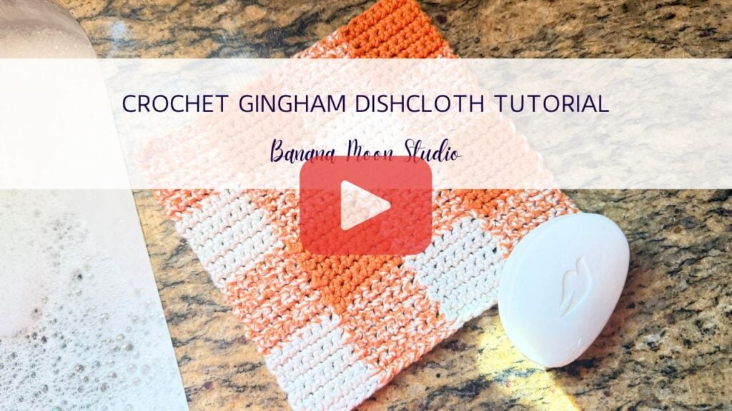 Crochet gingham dishcloth on a granite countertop with a bar of soap. A banner with text reads: Crochet gingham dishcloth tutorial, banana moon studio. A red and white play button is centered on the image.