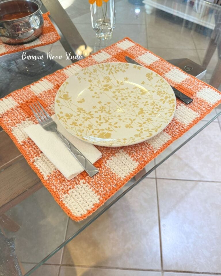 Orange and white crochet gingham placemat pattern lying flat on a glass table top set with plate, utensils, and napkin. Text on the image reads: Banana Moon Studio.