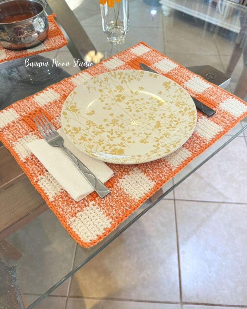 Orange and white crochet gingham placemat pattern lying flat on a glass table top set with plate, utensils, and napkin. Text on the image reads: Banana Moon Studio.
